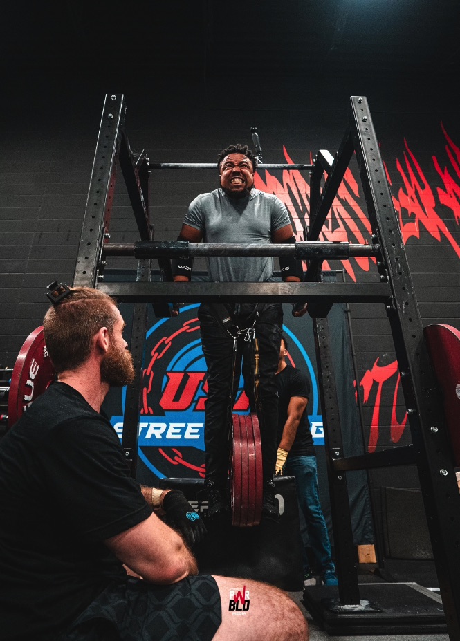 Nick attempts a weighted dip with loaded plates beneath him while spotters watch.